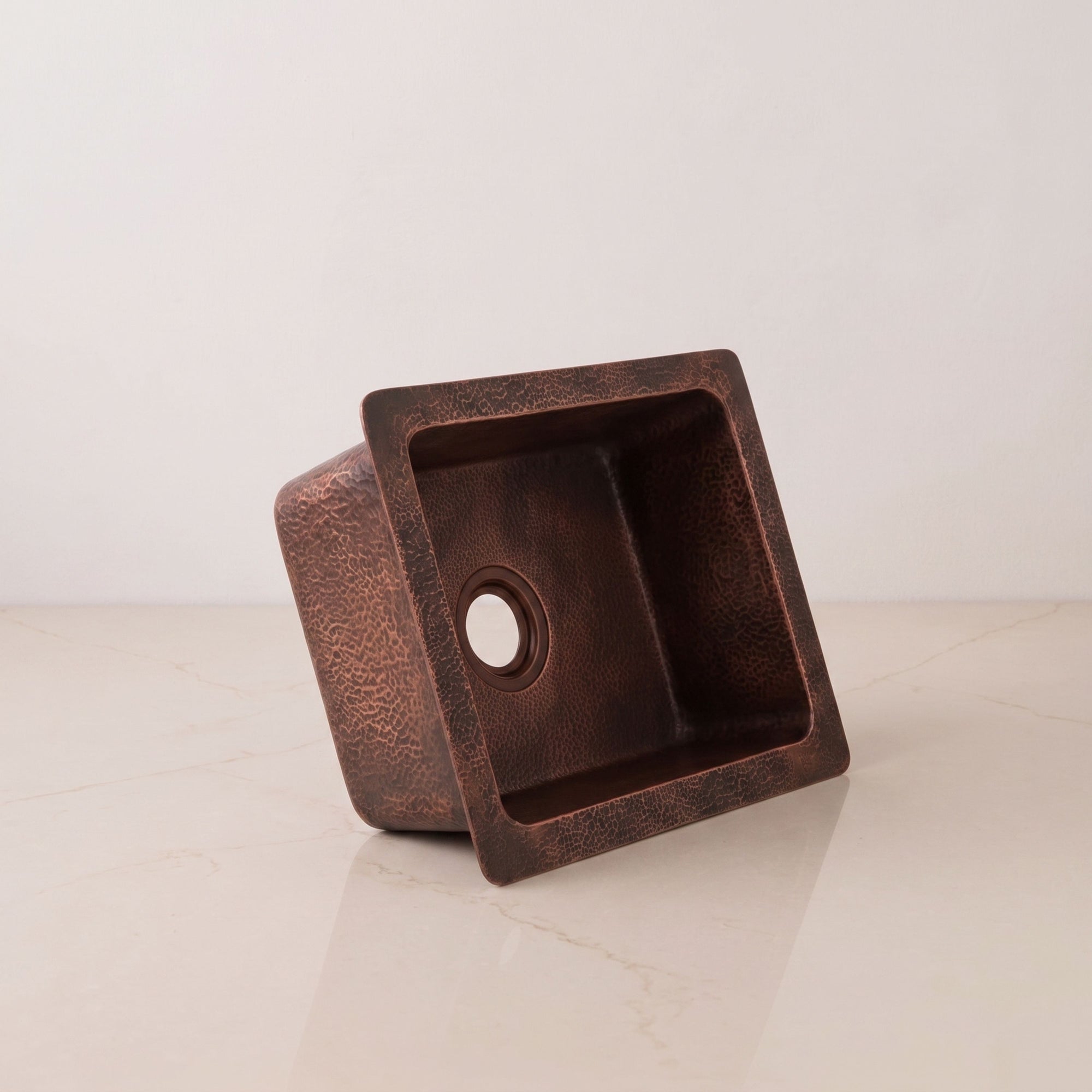 A single-square copper kitchen sink with a textured, hammered finish in a warm bronze-brown color, shown on a light beige marble surface against a neutral off-white background.