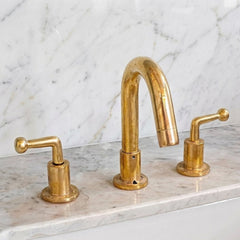 A three-hole bathroom faucet set in polished brass with a central arched spout and two matching lever handles, installed on a white marble countertop with a matching marble backsplash.