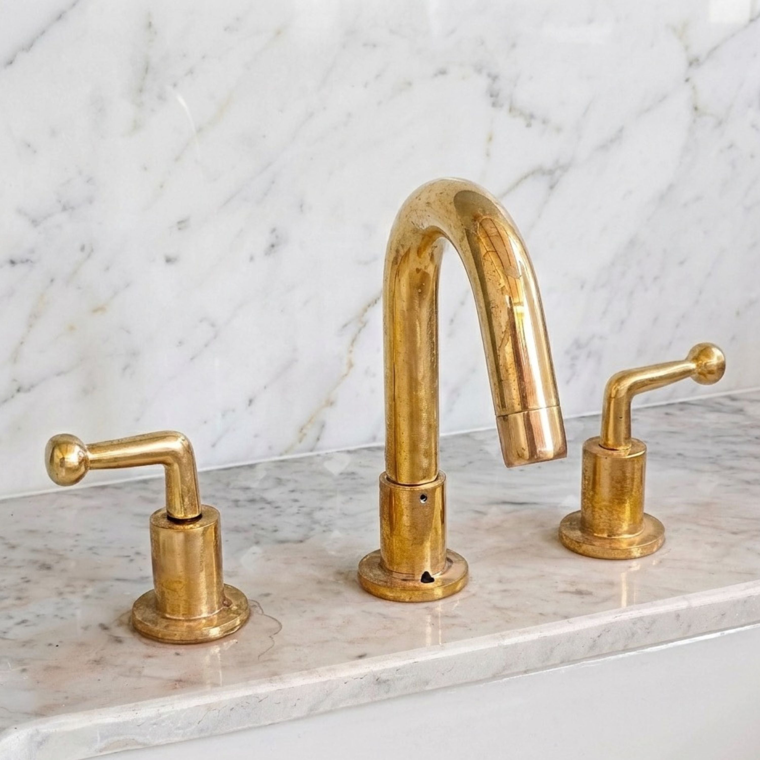 A three-hole bathroom faucet set in polished brass with a central arched spout and two matching lever handles, installed on a white marble countertop with a matching marble backsplash.