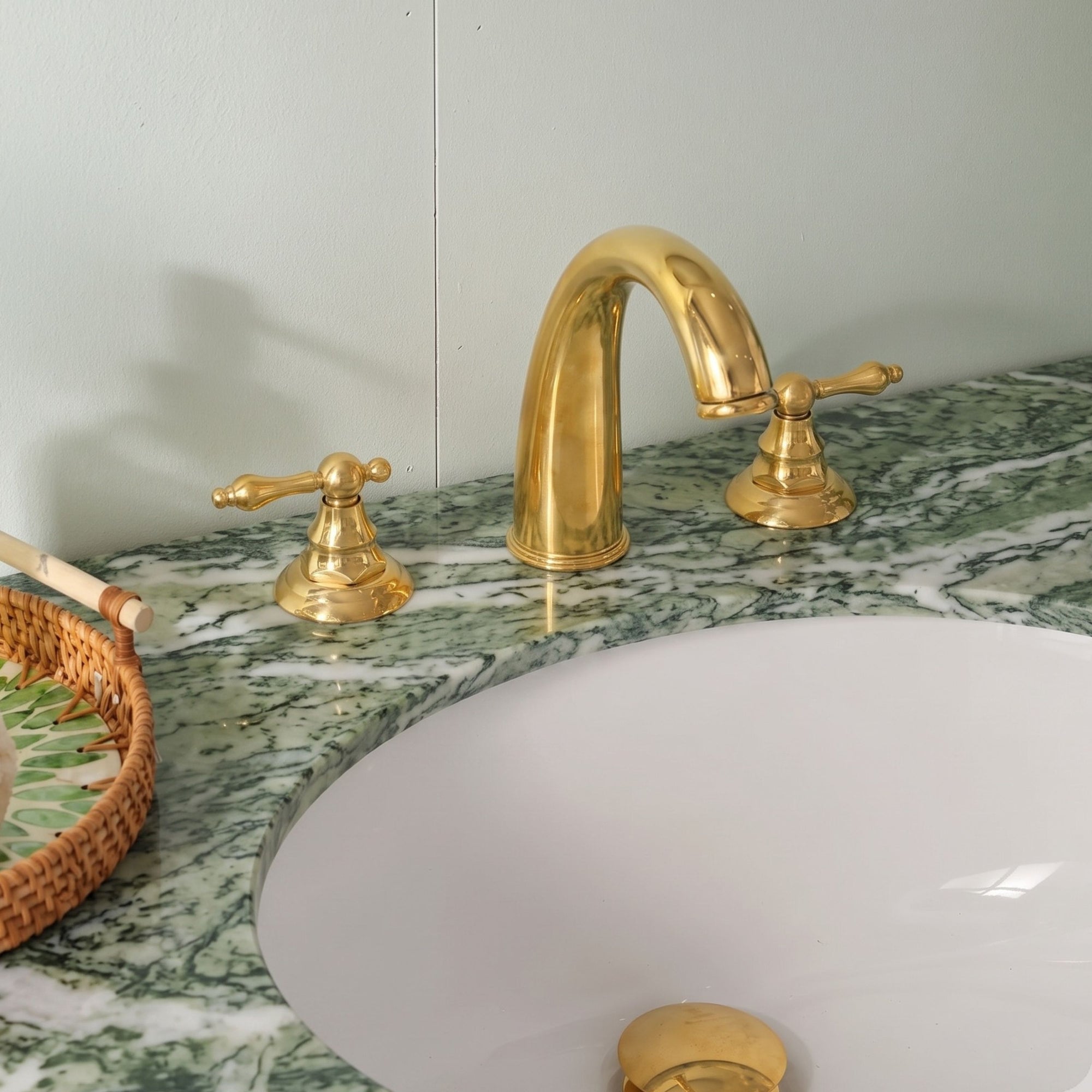 A polished gold brass bathroom faucet set with a curved spout and two separate handles, installed on a green and white marble countertop next to a white sink basin.