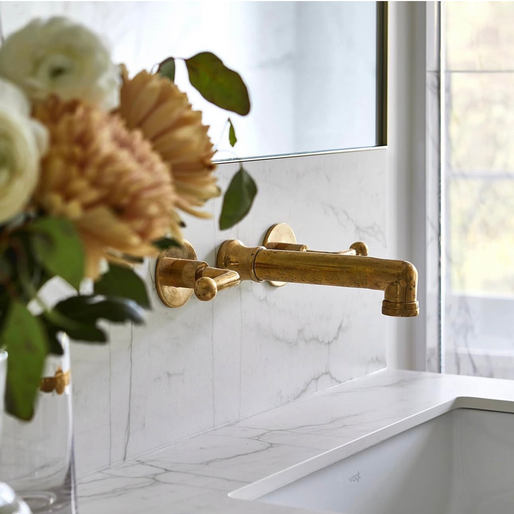 A close-up of a matte brass wall-mounted faucet installed above a white marble sink, with dried flowers in the foreground and a window in the background.