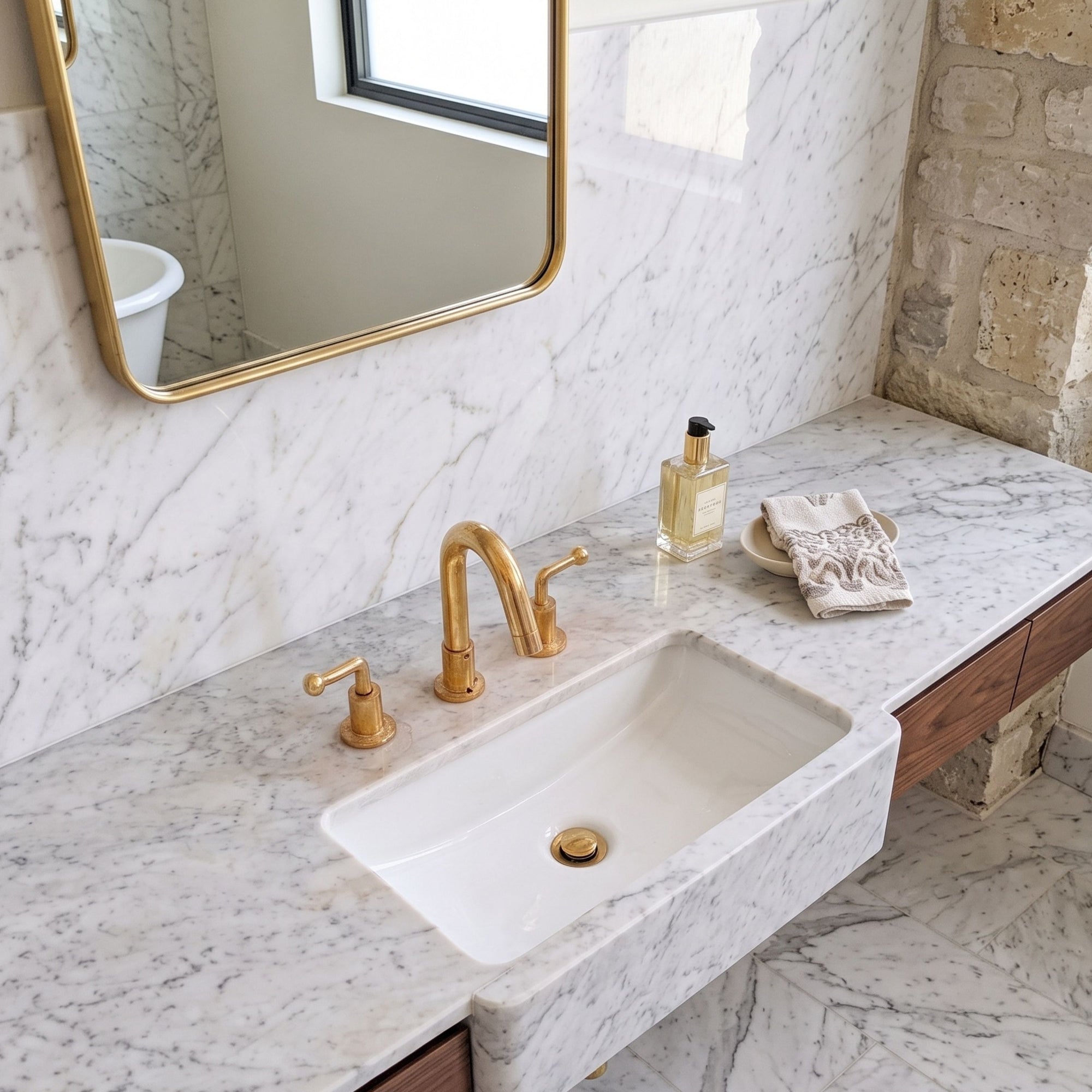A modern bathroom vanity featuring a white rectangular stone sink set into a white marble countertop with gold-finished faucet and handles, a gold-framed mirror, and a small decorative bottle and towel on the counter.
