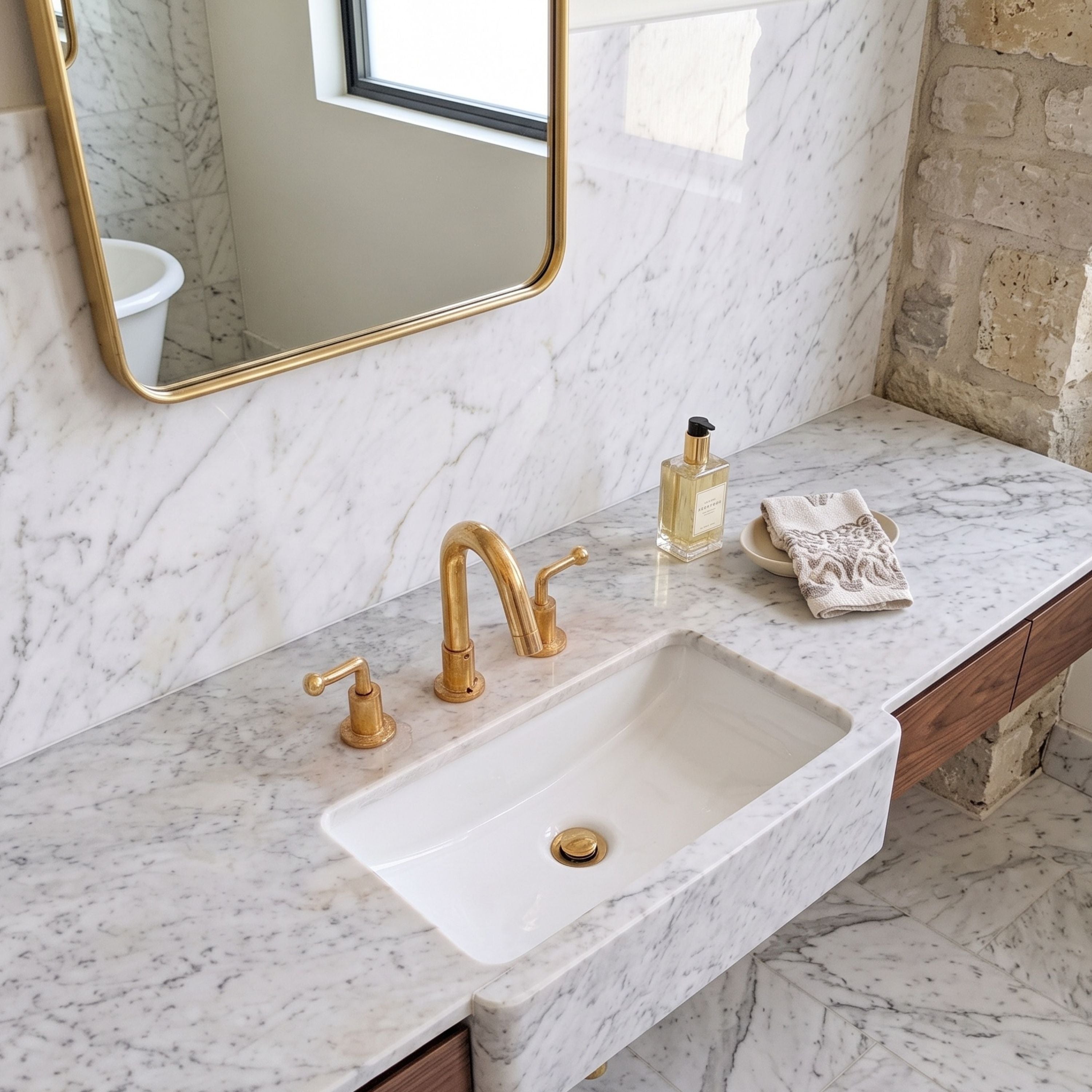 A modern bathroom vanity featuring a white rectangular stone sink set into a white marble countertop with gold-finished faucet and handles, a gold-framed mirror, and a small decorative bottle and towel on the counter.