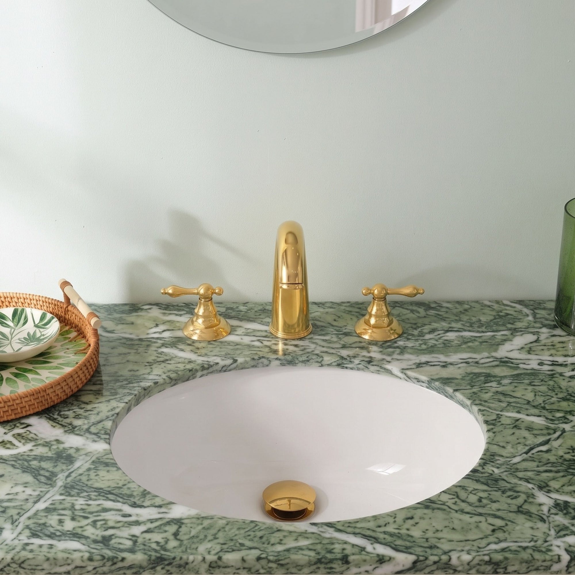 A polished gold faucet set with two handles and a central spout installed above a white oval sink, set into a green and white veined marble countertop.