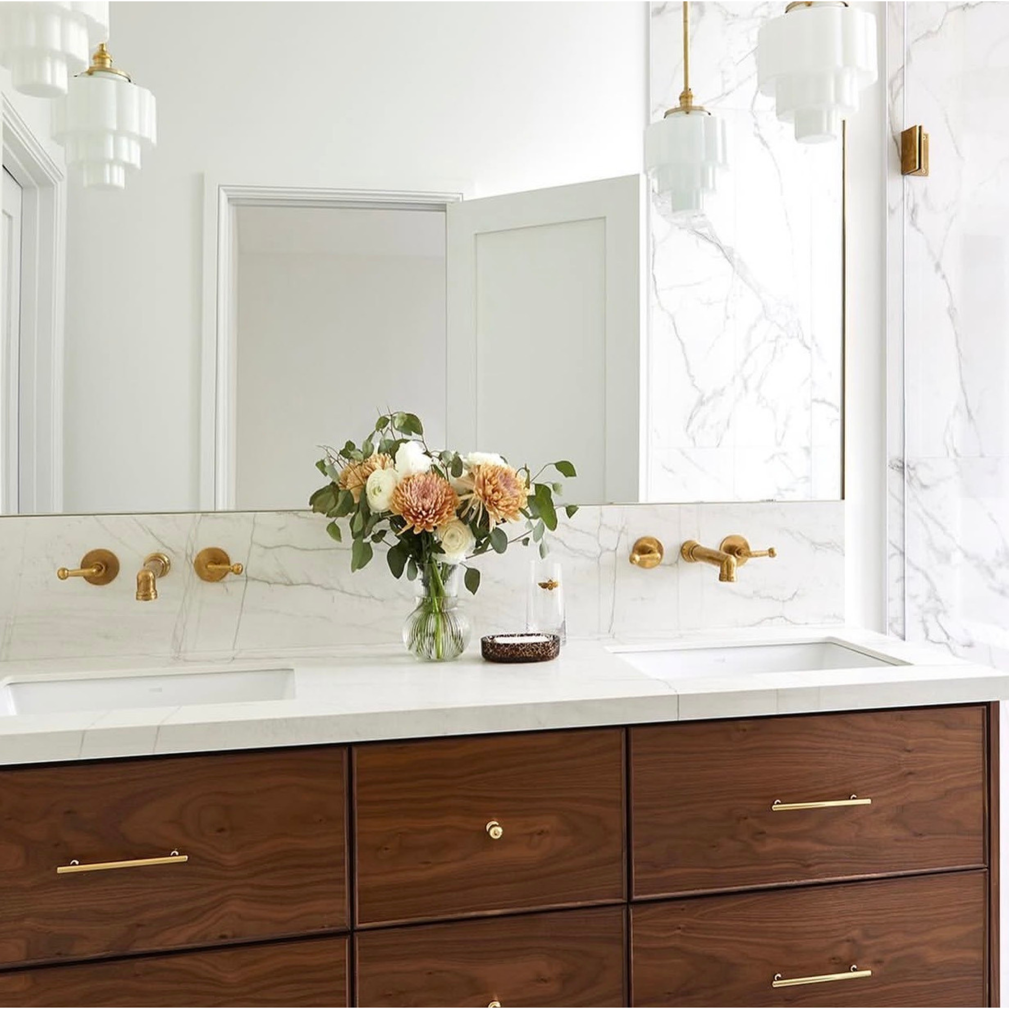 A modern bathroom vanity with a white marble countertop and backsplash, dark wood cabinets with brass handles, a large mirror, and pendant lights. A vase of flowers sits on the counter.