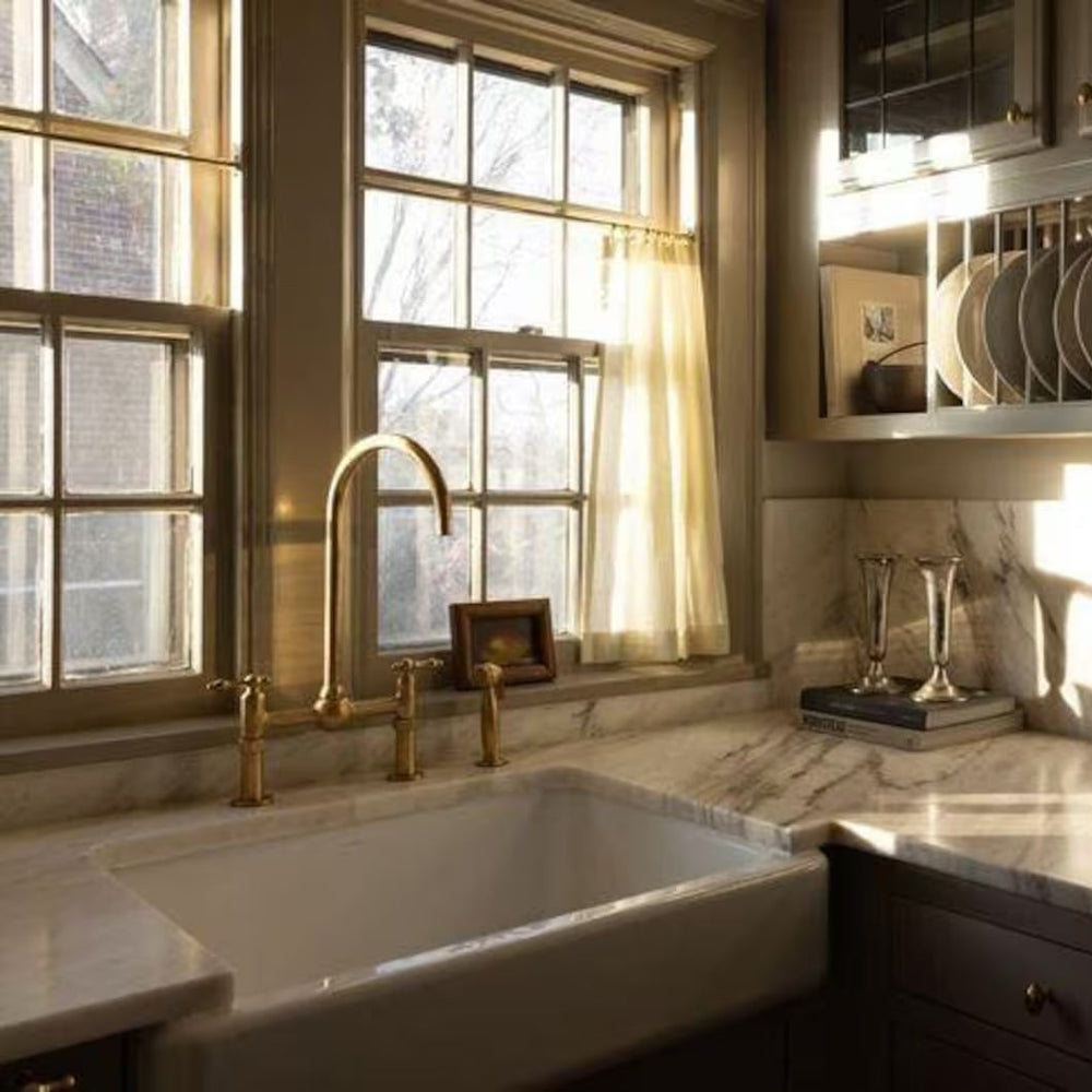 A kitchen setting showing an aged un lacquered brass faucet installed on a sink, with light reflecting off the surface and surrounding cabinetry.
