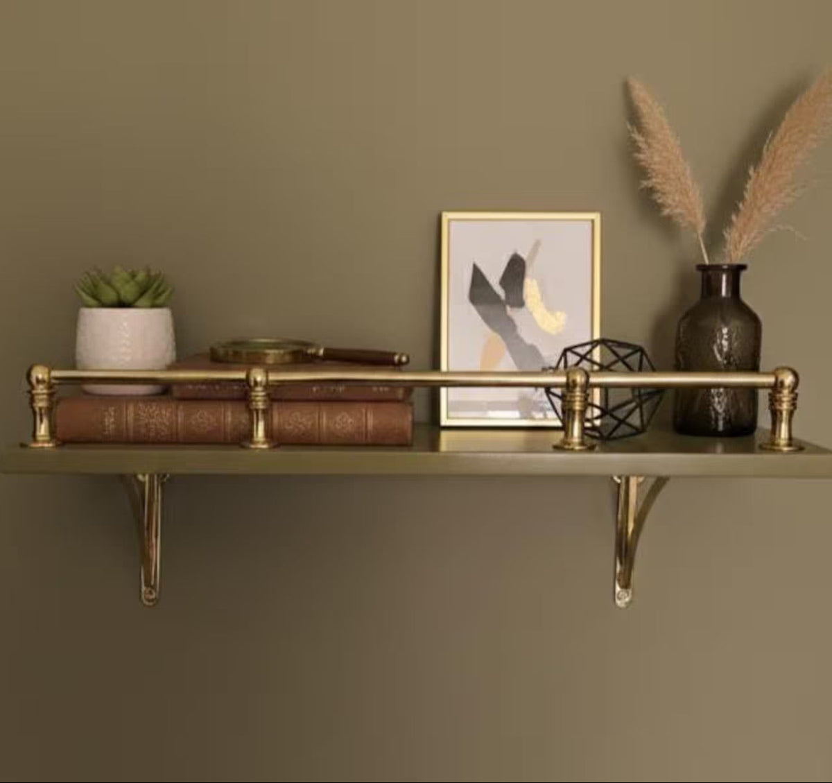 A wall-mounted shelf with a brass-toned metal frame and two gold brackets, styled with books, a small potted succulent, a framed print, a black geometric decor object, and a dark vase with pampas grass against a muted olive-gray wall.