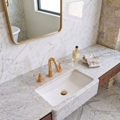 A modern bathroom vanity featuring a white rectangular stone sink set into a white marble countertop with gold-finished faucet and handles, a gold-framed mirror, and a small decorative bottle and towel on the counter.