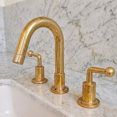 A polished brass three-hole bathroom faucet set with a high-arc spout, a side lever, and a separate soap dispenser handle, installed on a white marble countertop with a matching marble backsplash.