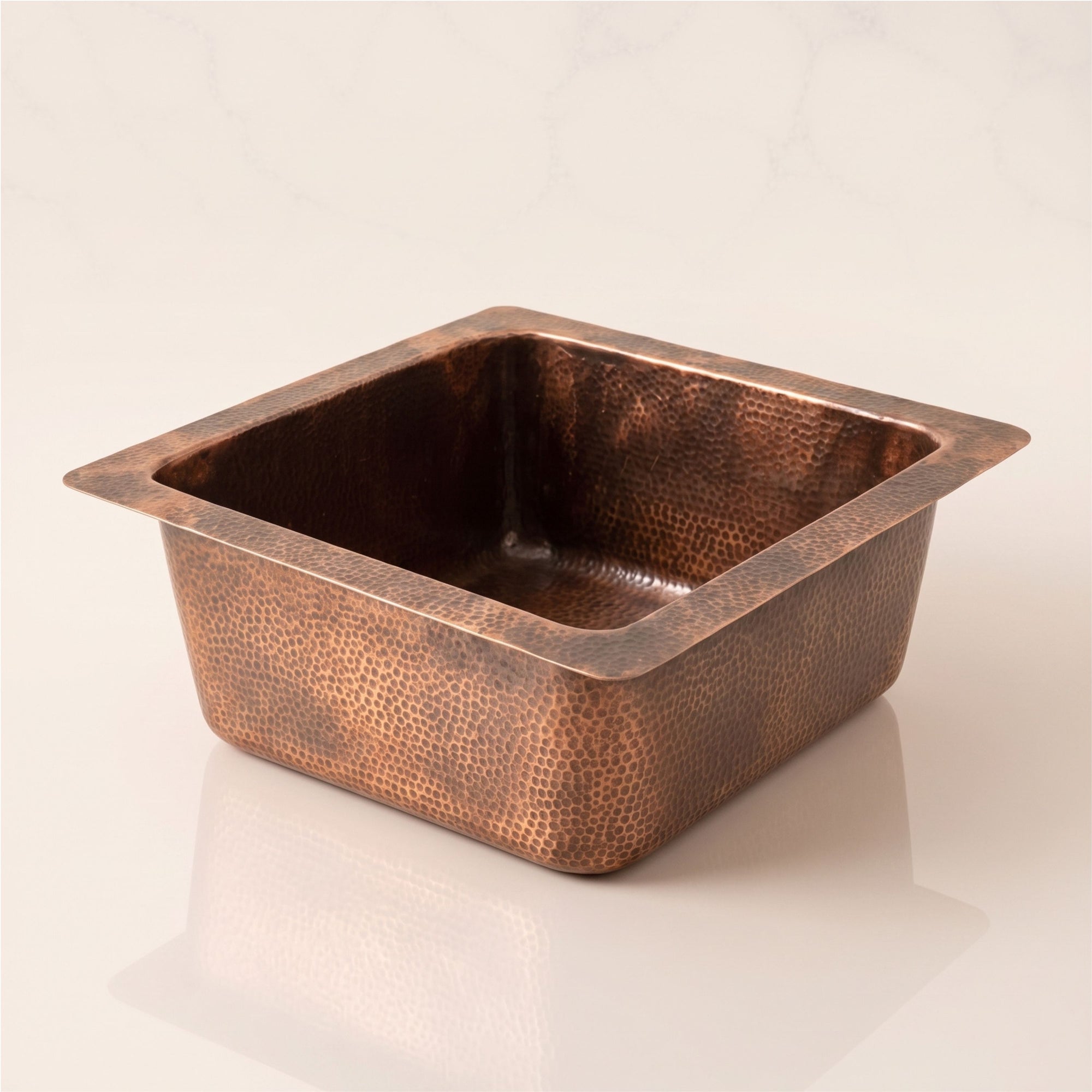 Close-up angled view of a handcrafted square solid copper bar sink with a hammered texture, resting on a polished marble countertop with its reflection visible.