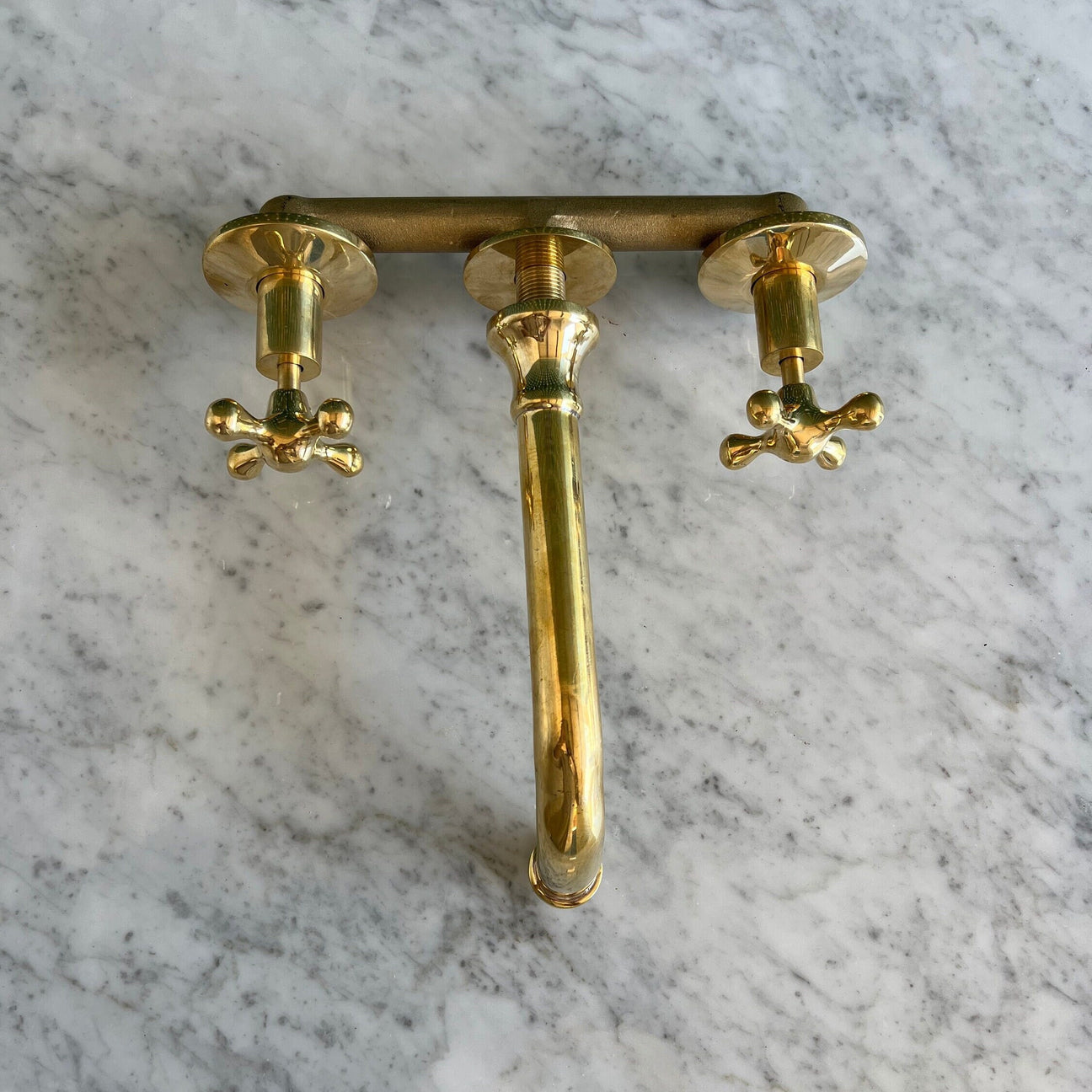 A polished brass three-hole faucet set with a central spout and two cross-handle valves, mounted on a white and gray veined marble surface.