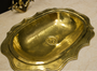 A vintage-style oval brass sink basin with a scalloped edge and a central drain, shown on a light beige marble countertop.
