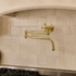 A wall-mounted kitchen faucet with two curved brass-style handles and a curved spout, installed against a beige tiled backsplash above a gas stove.