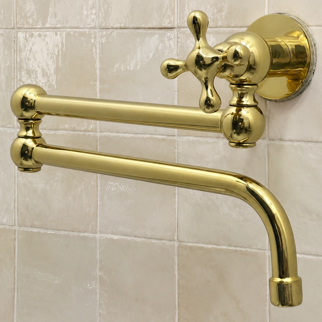 A polished gold wall-mounted faucet with a cross handle and curved spout, installed against a beige tiled wall.